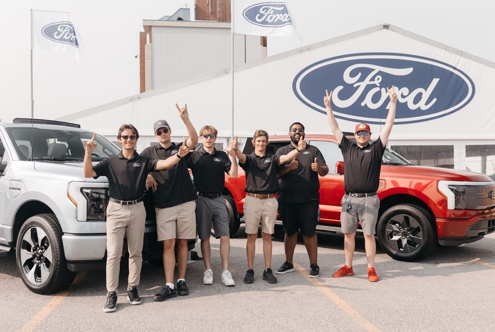 Sáu người mặc áo sơ mi đen giống nhau đứng trước một căn lều có dòng chữ "Ford" và hai chiếc xe.