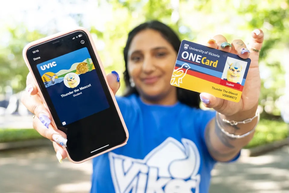 Staff member in blue UVic Vikes shirt holds up UVic One Card in one hand and mobile phone with One Card app in other.