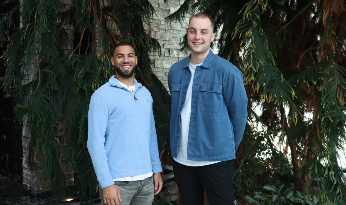 Two smiling young people standing beside each other against a backdrop of greenery.